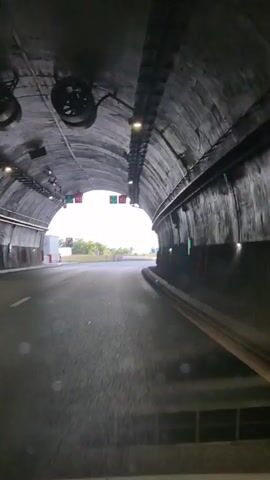 A first-person perspective video shows a car driving through a tunnel and then emerging onto an open road. The tunnel has concrete walls and a curved ceiling with lights. Signs are visible on the walls and overhead. Outside the tunnel, the road continues with lanes marked by white lines, and guardrails line the sides. Streetlights are also visible along the road. The sky is cloudy, and distant hills can be seen.