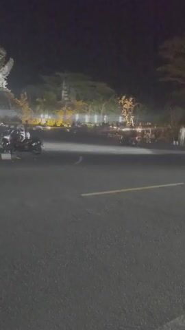 At night, a dark-colored car performs a controlled drift around a roundabout. There are several scooters parked to the left of the camera, and a group of people stand watching from the side of the road.  The roundabout has a statue in the middle and appears to be landscaped with lit trees and shrubbery.