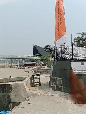 The video shows a sandy beach with several fishing boats pulled ashore. In the background, a white pier extends over the water, and a large turtle sculpture is visible near a building. The sky is overcast. The camera pans from right to left, revealing more of the beach and the ocean. There are no people clearly visible in the video, and the overall impression is one of a quiet, somewhat neglected coastal area.