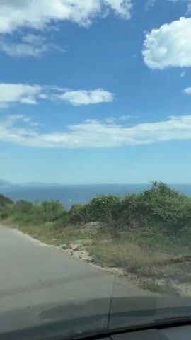 A car drives along a paved road. The road borders short, green vegetation. The view from the road reveals a body of water, and the sky is a light blue with several fluffy, white clouds.