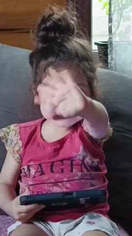 A young girl with curly brown hair tied in a messy bun is seated indoors, likely on a sofa, and facing the camera. She has bright eyes and a cheerful expression, smiling broadly. She is wearing a short-sleeved pink t-shirt with floral patterns and a partial visible text that seems to be "MAGINE" (possibly part of "IMAGINE"), paired with white shorts with a faint pattern.

In her left hand, she holds a dark-colored smartphone or small tablet. With her right hand, she waves playfully towards the camera, extending her fingers. The background shows a comfortable indoor setting, with a dark sofa behind her and a window to the right, through which some green foliage is visible, suggesting natural light.

The girl speaks in Russian, saying "красить" (to paint) and then "надо себя красить" (need to paint myself), with a joyful tone. Her demeanor is lively and engaging. The scene takes place in Barda, Russia.