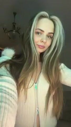 A woman is centered in the frame, recording a video of herself. She has long, straight hair with blonde streaks and light eyes. She is wearing a white, fuzzy cardigan sweater with a zipper down the front. The sweater has a furry hood. The background is a dimly lit room with a chandelier visible in the upper left corner. The woman looks into the camera, moves slightly, and smiles.