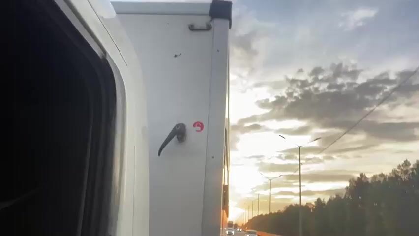 A close-up, side view of a white truck cab, with the passenger door ajar, reveals a black interior. The truck is on a highway during what appears to be sunset. The sun is low on the horizon, casting a warm glow through a cloudy sky. Streetlights are visible along the highway, and trees line the road. Traffic is moving on the highway, with several cars visible. The overall scene suggests a journey in progress during the golden hour.