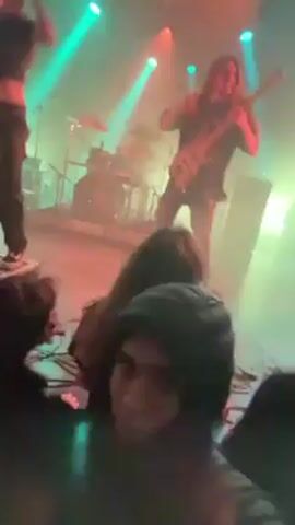 A video taken from a low angle shows a musical performance with a lead singer on a speaker.  A band is playing behind her.  The audience is crowded in front of the stage.  Red and green stage lights illuminate the scene, with smoke visible in the air.  A person in the foreground wears a black beanie and looks up at the stage.  At least one audience member in the foreground raises their hand with devil horn fingers.