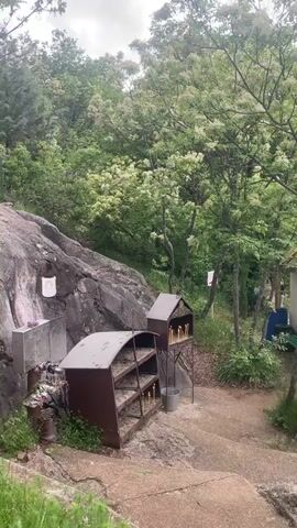 The video shows an outdoor scene at a religious site. In the foreground are metal candle stands with lit candles and a bucket. To the left, there is a large rock with religious icons. The ground is a mix of concrete steps and dirt pathways. In the background, there is a small white building with an open door where people are entering. A tree laden with white blossoms partially obscures the building. To the right of the building is a pole with a cross at the top and a Bulgarian flag hanging below. People are walking towards the building. One person carries a bundle of white fabric. The overall atmosphere is serene.