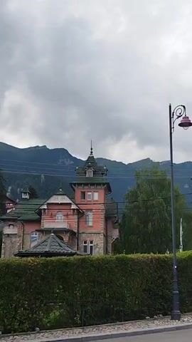 This is a vertical video filmed from a moving vehicle on a cloudy day. The camera pans to the right, revealing a picturesque European village nestled against a backdrop of majestic mountains. In the foreground, a neatly trimmed hedge lines the sidewalk, with a vintage-style lamppost standing tall beside it. Beyond the hedge, a charming pink house with a green roof and a tower draws the eye. Further down the street, several other quaint houses with distinctive architectural styles, including striped facades and steeply pitched roofs, are visible. A few people are seen walking along the street, adding a touch of life to the serene scene. The lush greenery of trees and the imposing mountain range complete the idyllic setting.