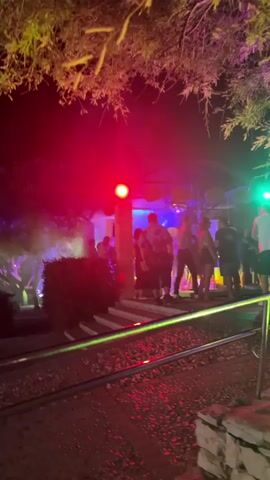 A group of people are gathered outdoors at night. The scene is lit by red and green lights. Some people are dancing, and others are standing around talking. There are trees and bushes visible in the background. The ground appears to be paved with stones. A railing is visible in the foreground.