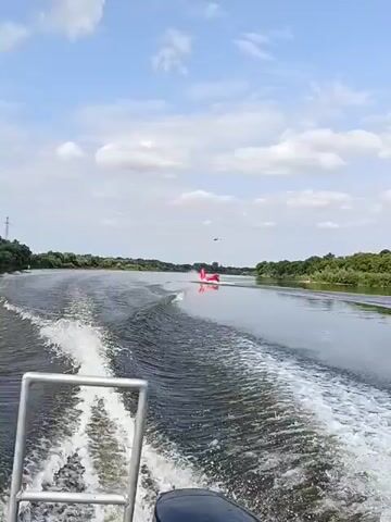The video shows a view from the back of a boat moving through a river. The boat is creating a large wake behind it. In the distance, another object, possibly another boat or a buoy, is visible on the right side of the river. The riverbanks are lined with green trees. The sky is blue with some white clouds. The scene appears to be during the daytime.