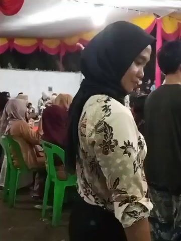 The video shows a gathering of people under a tent with white and pink draping. In the foreground, a woman wearing a black hijab and a patterned shirt is visible from the side and then turns to face the camera briefly. She is looking to her right. Behind her, other attendees are seated at tables. The environment appears to be outdoors at night, indicated by the darkness beyond the lit tent. The people are dressed in casual to semi-formal attire. Some are wearing hijabs. A man in a patterned shirt and cap is seen in the background to the right. There are green plastic chairs and tables.