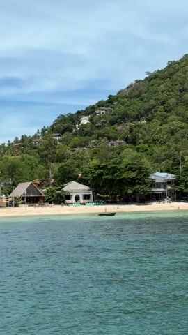 A video shows a view of a tropical island with a lush green mountain slope covered in trees.  Several buildings, including what appears to be a resort or houses, are scattered along the hillside. In the foreground, there is a sandy beach with a few people.  The ocean in front of the beach is a clear turquoise color, and there is a small dark boat floating on the water. Several people can be seen swimming or snorkeling in the ocean near the beach. The sky is blue with scattered white clouds.