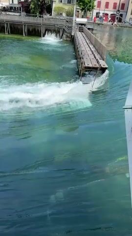 The video shows a river with turbulent water flowing past a wooden structure that appears to be a bridge or dam. The water is a striking two-toned blue and green, with the green section near the structure being much more frothy and turbulent. In the background, there are buildings and trees lining the riverbank. The sky is clear and blue with some clouds. No people or animals are clearly visible in the video. The overall scene depicts a natural landscape with man-made elements, highlighting the power and beauty of the flowing water.