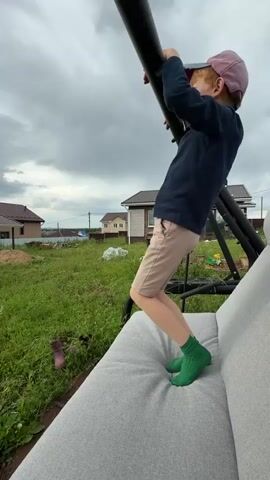 This video features a young boy with red hair, wearing a dark blue hoodie, light beige shorts, and bright green socks. He is standing on a gray cushion, holding onto a black metal bar, appearing to look through something like a telescope or binoculars. He is wearing a pink baseball cap.

The scene is outdoors in a rural or suburban setting, likely in Pyachanki, Russia. In the background, there are houses with reddish-brown roofs and one with a light-colored facade. The area is covered in green grass and some construction materials, suggesting a backyard or a newly developed area. The sky is overcast and cloudy, indicating a possibly dull or stormy day.

The boy seems engrossed in what he is observing, and his posture suggests a sense of focus or curiosity. The spoken Russian phrase "Была бы крыша, был бы кайф" translates to "If there were a roof, it would be a blast," which might imply his desire for a more sheltered or playful environment, or perhaps a humorous observation related to the weather or his current activity. The overall mood is casual and playful, capturing a moment of childhood exploration.