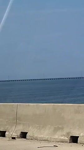 The camera pans across a long, straight bridge spanning a wide body of water under a partly cloudy sky. The bridge appears to be supported by many pilings, and there are some structures in the water to the right of the frame. A low concrete barrier is visible in the foreground, suggesting the video was shot from a vehicle.