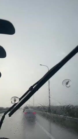 The video shows a view from inside a car driving on a highway during a rainstorm. The windshield is covered in raindrops, and a windshield wiper is visible at the bottom of the frame. Several cars are ahead on the wet road, their brake lights illuminated. Streetlights and vegetation line the sides of the highway. The sky is overcast and grey.