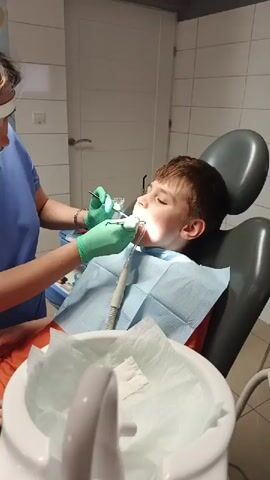 A young boy sits in a dental chair, his mouth open as a dentist in green gloves works on his teeth. The dentist is using dental tools, including a dental mirror and a probe, to examine and treat the boy's teeth. A dental suction tube is positioned in the boy's mouth to remove saliva. The environment appears to be a dental operatory room, with tiled walls and a dental light illuminating the boy's face. A tray of dental instruments and supplies is visible on the left side of the frame. The boy is wearing a blue dental bib.
