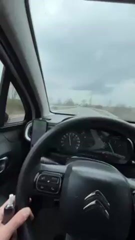 Here is a detailed description of the video:

The video is shot from inside a car, focusing on the dashboard, steering wheel, and the road ahead. The dashboard is predominantly black, with a speedometer showing the car traveling at around 93 km/h. A smartphone is mounted on the dashboard, presumably for navigation. The driver’s hands are visible on the steering wheel, and in one hand, they are holding a small, gray, cylindrical object.

Outside the car, the road stretches into the distance under a cloudy sky. Trees line the sides of the road. Another vehicle can be seen ahead in the same lane. The scene gives the impression of a drive on a highway or major road during overcast weather. A rap song plays in the background.