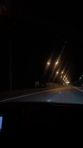 The video shows a nighttime drive from the driver's perspective. The interior of the car is dimly lit, revealing the dashboard, steering wheel, and infotainment screen. The infotainment screen displays music playing. The car is traveling on a highway lined with streetlights. The road ahead is visible through the windshield, showing lane markings and the dark surroundings.