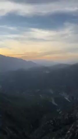 The video shows a scenic mountainous landscape at dusk. The camera pans to reveal a rocky hilltop with sparse vegetation and then focuses on two parked cars: a yellow convertible Porsche and a black BMW sedan. The cars are parked on a dirt or gravel surface near a steep, rocky hillside.