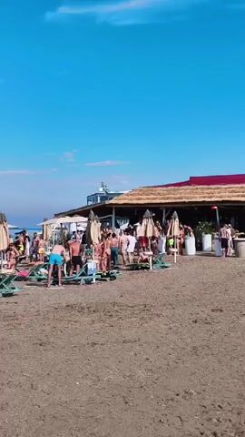 A beach scene on a clear sunny day. The sand is a light brown color. In the foreground, there are several empty lounge chairs and umbrellas. Further back, a group of people are scattered across the beach. Some are sitting on lounge chairs, while others are standing and interacting. There appears to be a beach bar or restaurant structure in the background with thatched roofing and tables. The ocean is visible behind the beach, with a clear blue sky above.