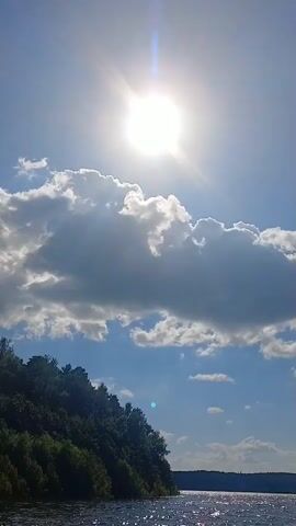 The video begins with a view of a bright sun shining through a partly cloudy sky over a large body of water. The sun's rays are visible, creating a halo effect. The water is calm and reflects the sunlight, creating a shimmering effect. On the left side of the frame, a lush green hillside with trees slopes down to the water's edge. As the camera pans to the right, the view opens up to a wider expanse of water with more distant hills in the background. A small boat can be seen in the distance. The camera continues to pan and moves forward, revealing a wooden pier extending out into the water. A Russian flag flies from a flagpole on the pier. Several people are seated on the pier, some of whom appear to be fishing. The scene suggests a peaceful day on the water, possibly a lake or a large river.