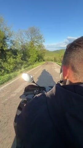 The video shows a person riding an ATV on a paved road through a rural area. The camera is mounted behind the rider, capturing their back and the view ahead. The road is cracked and has grass growing along the edges. On either side of the road, there are fields of green grass and wooded areas. The sky is blue with some clouds, and the sun is shining brightly.  The video shows a slightly tilted perspective, revealing a wide view of the rural landscape, as the ATV travels along the curving road. The rider is wearing a dark hoodie.