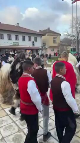 The video shows a group of people in what appears to be a traditional Bulgarian celebration. The setting is a paved square or street in a town, with buildings visible in the background. Some people are dressed in traditional costumes, including furry outfits with masks and vests with embroidered patterns. There are also people carrying the Bulgarian flag. The sound of bells can be heard, suggesting they are part of the celebration. The people are moving and dancing, creating a festive and lively atmosphere. The sky is overcast.