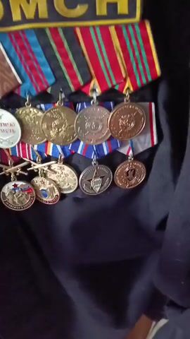 The video displays a close-up of a dark-colored uniform, likely military or law enforcement, adorned with numerous medals and ribbons. At the top of the uniform, a yellow patch with the letters "OMCH" (ОМЧ) is visible, along with a silver-colored emblem or badge above it. Below the patch, there are several rows of medals.

The medals are attached to colorful ribbons, featuring various patterns and colors including red, blue, green, gold, white, and brown. Some medals are gold-toned, while others are silver-toned. In the upper row of medals, a silver medal on the far left clearly shows the text "ЗА ВЕРНУЮ СЛУЖБУ", which translates to "FOR FAITHFUL SERVICE". In the lower row, a silver-toned medal prominently features the number "100". The details on other medals are not clearly discernible due to their small size and the video's resolution. The uniform's fabric and the metallic sheen of the medals are clearly visible. The background is a soft, out-of-focus dark fabric, keeping the focus entirely on the uniform and its decorations.
