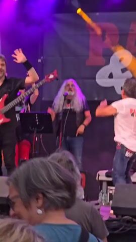 A low-angle shot captures a live outdoor concert scene. Several male musicians are visible on stage. One man with long grey hair and a black vest holds a microphone and gestures with his right hand. To his right, another man in a white t-shirt and jeans turns away from the camera, also gesturing with his right hand. Behind them, a drummer with long dark hair vigorously plays a Roland drum kit, lifting a drumstick high in the air. To the drummer's right, a guitarist with dark, shoulder-length hair and glasses, wearing a black jacket with gold detailing, plays an electric guitar. In the foreground, the backs of several audience members' heads are visible, indicating their proximity to the stage. A large banner with text, including "LA COSA NOS," hangs behind the musicians. Bright stage lights illuminate the scene with orange and purple hues.
