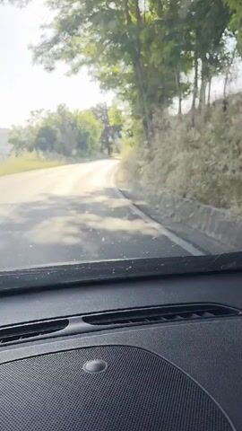 The video shows a first-person perspective from inside a car driving down a paved road. The road is bordered by green trees and vegetation on both sides, with a slight incline to the right. Sunlight streams down, creating some glare on the windshield, which has a few water droplets. The car's dashboard is visible in the foreground, including a speaker grill and a vent. The road ahead curves slightly to the left and then straightens. The sky is clear and bright.