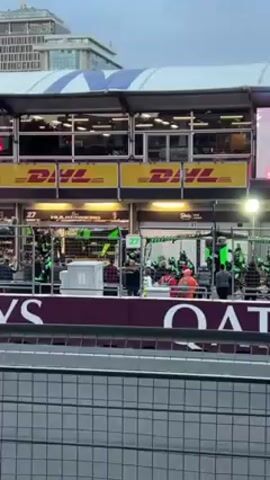 The video shows a Formula 1 race pit stop. There are several pit crew members in green shirts and helmets working on a car. Above the pit row is a large screen showing replays of the race. The pit lane is bordered by a low wall with the word "QATAR" printed on it. The weather appears overcast.