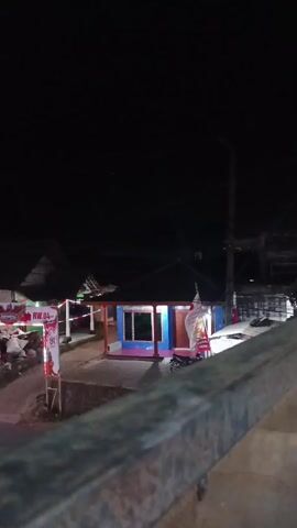 A vertical, nighttime, low-angle shot captures a small, brightly lit building with blue and pink accents, situated in what appears to be a rural or suburban environment. A dark, starless sky dominates the upper portion of the frame. In the foreground, a rusty, metallic railing runs horizontally across the bottom, slightly out of focus. To the left of the building, a banner with red and white stripes and text that appears to read "RW 04" is visible, along with a pile of what looks like discarded items. The building's windows are illuminated, casting a warm glow. A tall pole, possibly a streetlight or utility pole, stands to the right of the building. The overall scene is dimly lit, with the building being the primary source of light. There is no discernible movement or interaction between people or objects within the frame.