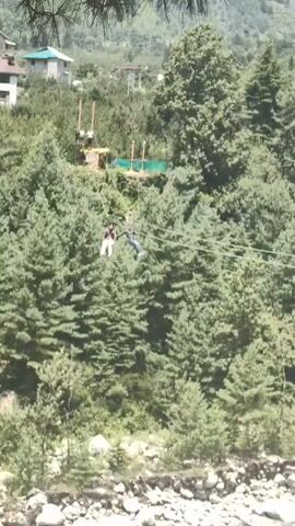 The video shows a person zip-lining through a forest with a river in the foreground. The zip-line appears to be suspended between two trees or poles, and the person is wearing a harness and helmet. The forest is dense with tall green trees, and the river is rocky and shallow. In the background, there are mountains and some buildings. The sky is blue with some clouds.