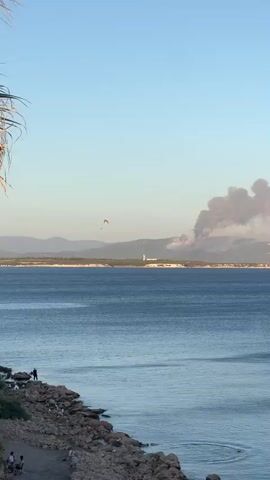 The video shows a wide shot of a body of water with a rocky shoreline in the foreground. On the far side of the water, a distant hill is visible with a large plume of smoke rising from it, indicating a fire. A small paraglider can be seen in the sky above the water, near the left side of the frame. A white lighthouse-like structure is visible on the hill, as is a red flag. The sky is clear and blue. The overall impression is one of a scenic landscape marred by a distant environmental event.