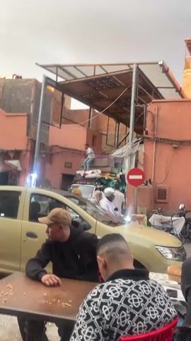 The video shows a street scene in a Middle Eastern or North African locale. The camera is held from the ground and shows a low-rise commercial street with pedestrians and traffic. In the foreground, two men sit at a small table near a light yellow car. The man on the left is wearing a brown baseball cap and a black hooded jacket. A second man in a patterned sweater is seen from behind. In the background, a small white truck is piled high with mattresses, luggage, and household goods. A man in light-colored jeans and a white shirt sits atop the pile, using a rope to secure the load. Other pedestrians are walking by. The buildings are constructed of brick and painted in light colors such as pink and yellow. A metal structure partially covers the street above. A red and white "do not enter" sign is visible.