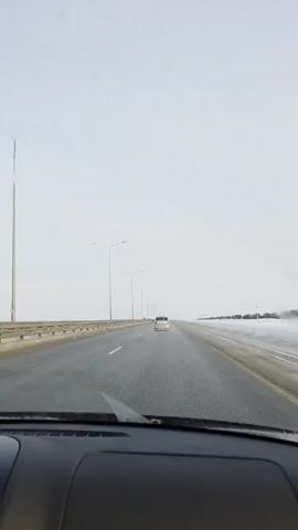 The video depicts a car driving on a highway under overcast, possibly snowy conditions. The wipers are active, clearing the windshield. The road is wet, and snow is visible on the roadside. Streetlights line the highway. A white vehicle is seen ahead in the same lane. The view is from inside the car, showing the dashboard. Music is audible.