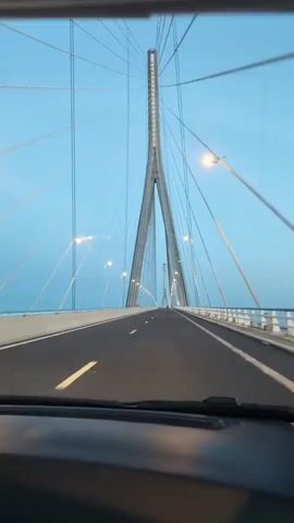 The video shows a view from a vehicle driving across a large, modern cable-stayed bridge. The sky is a clear, pale blue, transitioning to soft hues of pink and orange near the horizon, suggesting either sunrise or sunset. The bridge's distinctive tall pylons and numerous cables are visible, stretching towards the sky. Road markings are clear on the asphalt. To the side, a body of water, possibly the sea or a large river, is visible. There are also faint outlines of industrial structures or distant buildings on the horizon. The video captures the smooth movement of the vehicle across the bridge, with the landscape gradually shifting in the background.