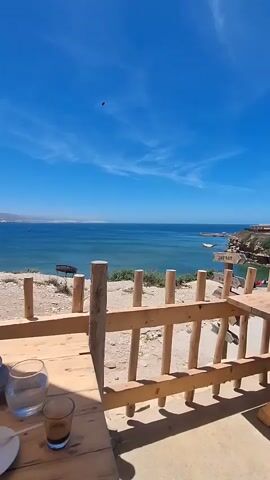 The video shows a scenic view of the ocean from a wooden deck. A wooden table and chairs are in the foreground, with drinks on the table. The ocean is a deep blue, and the sky is a bright blue with scattered white clouds. There are some buildings and cliffs visible in the distance. The overall impression is one of a relaxing outdoor setting with a beautiful view.