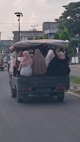 A group of women wearing hijabs are riding in the back of a pickup truck on a road. The truck has a canopy covering the bed. Buildings and a street light are visible in the background. The truck has a license plate that reads "BL 8416 BA". The sky is cloudy.
