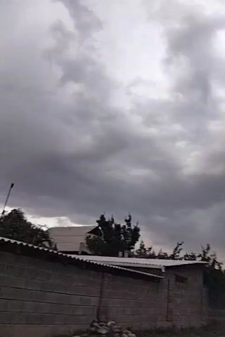 A video shows a grey cloudy sky over a brick wall with a corrugated roof. Trees are visible behind the wall. The camera pans to the right, showing a dirt road.