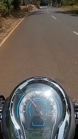 This is a first-person perspective video from a motorcycle. The motorcycle is traveling on a paved road. The dashboard of the motorcycle is visible in the foreground, showing the speedometer, an odometer reading of 25636 kilometers, and a fuel gauge. The road ahead is winding and lined with trees and vegetation. There are a few other motorcycles visible in the distance on the road. The weather appears to be sunny.