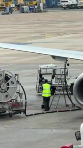 The video shows the nose of a red-painted airplane being refueled on an airport tarmac. The nose of the plane is painted red, with the letters "DYM" in white just above the cockpit windows. The plane has a large silver engine visible on its right side, and a fueling hose is connected to the plane's wing. A fuel truck is parked near the plane with a ladder leading to a point on the wing, and two ground crew workers in yellow safety vests are positioned near the plane and fuel truck. Other airport service vehicles are visible in the background.