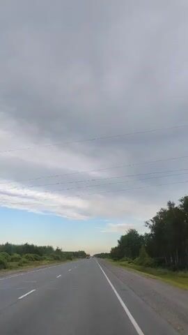 A vertical video shows a highway with a cloudy sky. The road is paved with asphalt and has white lane markings. On either side of the road are green trees and bushes. In the distance, several vehicles are visible, including a yellow truck driving towards the camera. The video captures a sense of open road and travel.