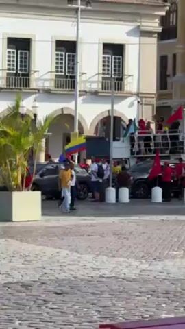 The video shows a public square with people milling about. In the center, several dark-colored cars are parked near a truck that appears to be a makeshift stage. People dressed in red shirts are standing on the top of the truck, waving flags, including the flags of Colombia and Brazil. Some people are standing around the vehicles, while others walk across the square.  The square is paved with cobblestones. The backdrop is a building that is predominantly white with architectural details, including arched porticos and shuttered windows. Another building with a yellow facade is partially visible.  The scene is bathed in sunlight.