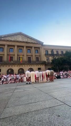A group of performers, mostly wearing white shirts and shorts, are in a line on a cobblestone plaza, bending over with their backs to the camera. One performer in the middle is performing a backflip over the line of bent-over individuals. A crowd of spectators is gathered behind the performers, sitting and standing. In the background, there is a large, ornate building with the words "DIPUTACION DE NAVARRA" written across its facade. Spanish flags are displayed on the building. The sky is clear and blue.