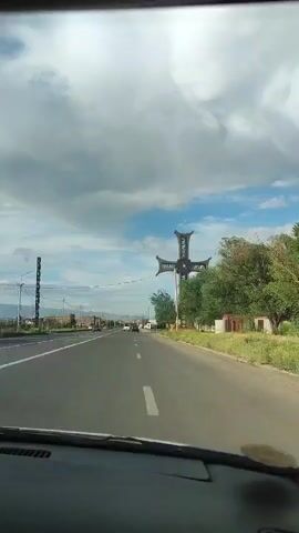 The video, captured from the passenger seat of a moving vehicle, showcases a large, ornate cross monument situated on the right side of a road. The cross is made of a dark material, possibly metal, with intricate designs and what appears to be a circular emblem in the center. The monument is quite tall and stands against a backdrop of a partly cloudy sky with patches of blue. Lush green trees and bushes are visible around the base of the cross and line the side of the road. In the distance, on the left side of the road, various buildings and structures can be seen, hinting at an urban or suburban environment. The road itself is paved and appears to have multiple lanes, with other vehicles intermittently visible in the distance. The overall impression is one of driving past a significant landmark in a moderately developed area under a pleasant sky.