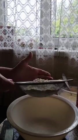 The video shows a close-up of a person's hands sifting flour into a large bowl. The flour is being sifted through a metal sieve, and a fine powder is falling into the bowl below. The background shows a window with white lace curtains, suggesting an indoor setting, likely a kitchen. The lighting is natural, coming from the window. There is also a glimpse of a digital scale under the bowl. The focus is on the action of sifting, with the hands and the sieve being the main subjects.