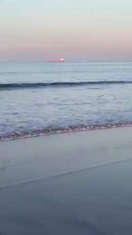 The video shows a beach with gentle waves lapping onto the sand. The water is calm and reflects the soft light of the sky, which is a mix of pale blues and pinks, indicating either sunrise or sunset. A ship can be seen in the distance at the beginning of the video.  Later, the silhouette of a coastal city is visible in the background. The beach itself is dark, likely due to the sand's composition.