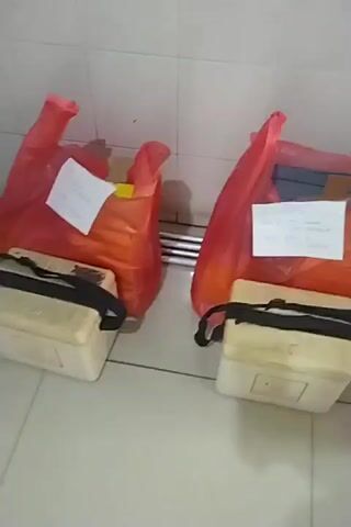 The video shows a row of items placed against a tiled wall. From left to right, there are two red plastic bags, each containing items and with a white paper note attached. In front of each red bag is a small, light-colored cooler bag with a black strap. Between the first two red bags is a blue-lidded cooler. To the right of the first cooler bag and in front of the second red bag is another cooler bag. Further to the right, there is a third red plastic bag with a yellow document and a white note, and a blue-lidded cooler container. The floor is tiled. The lighting appears to be artificial. The scene suggests a storage or waiting area, possibly for deliveries or distribution.