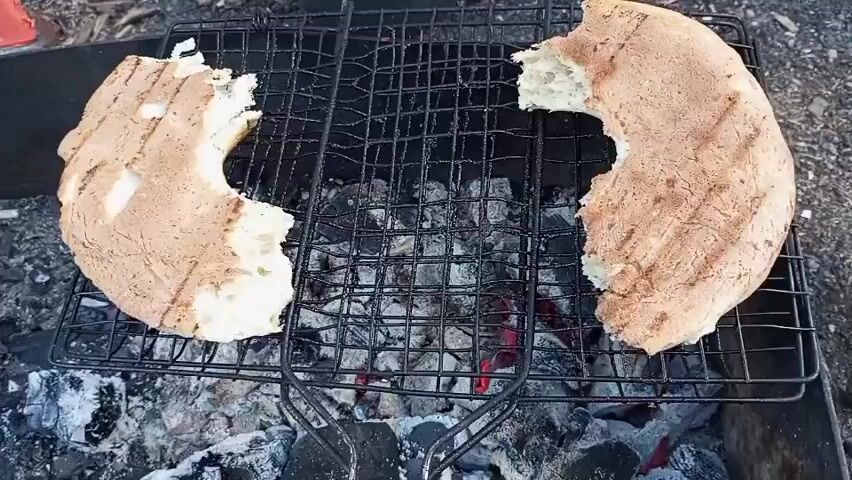 Two pieces of round flatbread with sections torn out are being grilled on a black metal grill rack over hot coals. The bread has grill marks. The background shows dark soil and some debris. Music plays.