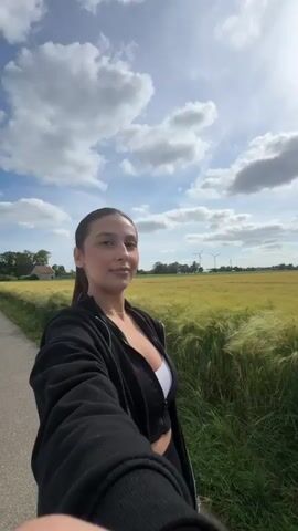 A young woman with dark hair in a ponytail, wearing a black jacket over a white top, stands on a paved path next to a field of crops. She first looks to her right, then turns to face the camera with a slight smile. In the background, there is a field, a partly cloudy sky, and distant wind turbines. The woman then moves out of the frame to the right. A white car's front bumper and hood are briefly visible as it passes from left to right on the path.